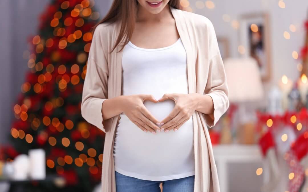 Pregnant lady making a heart with her hands over her belly with a Christmas tree in the background