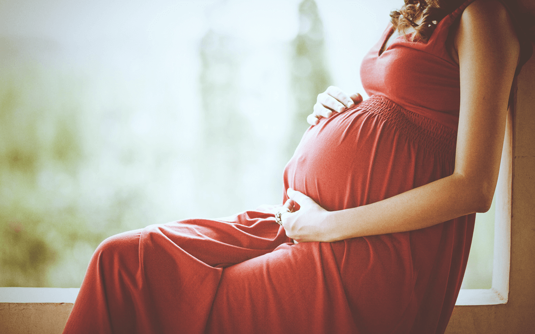 Pregnant lady in a red dress sat in a window holding her belly