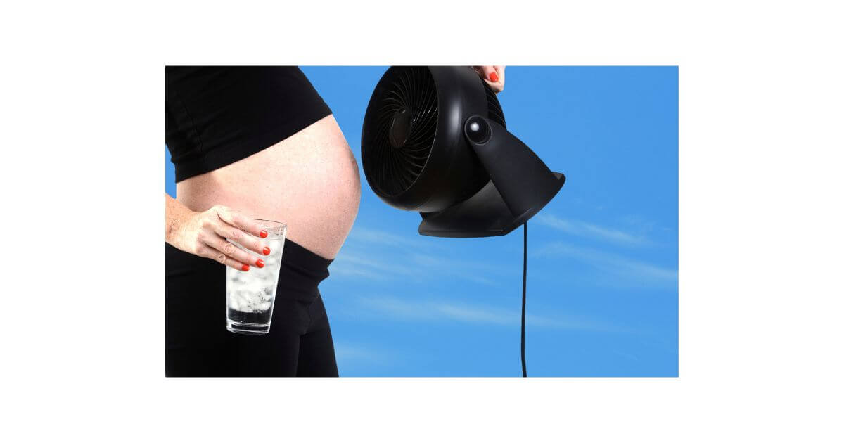 Pregnant woman wearing a black crop top holding a glass of iced water and a cooling fan.
