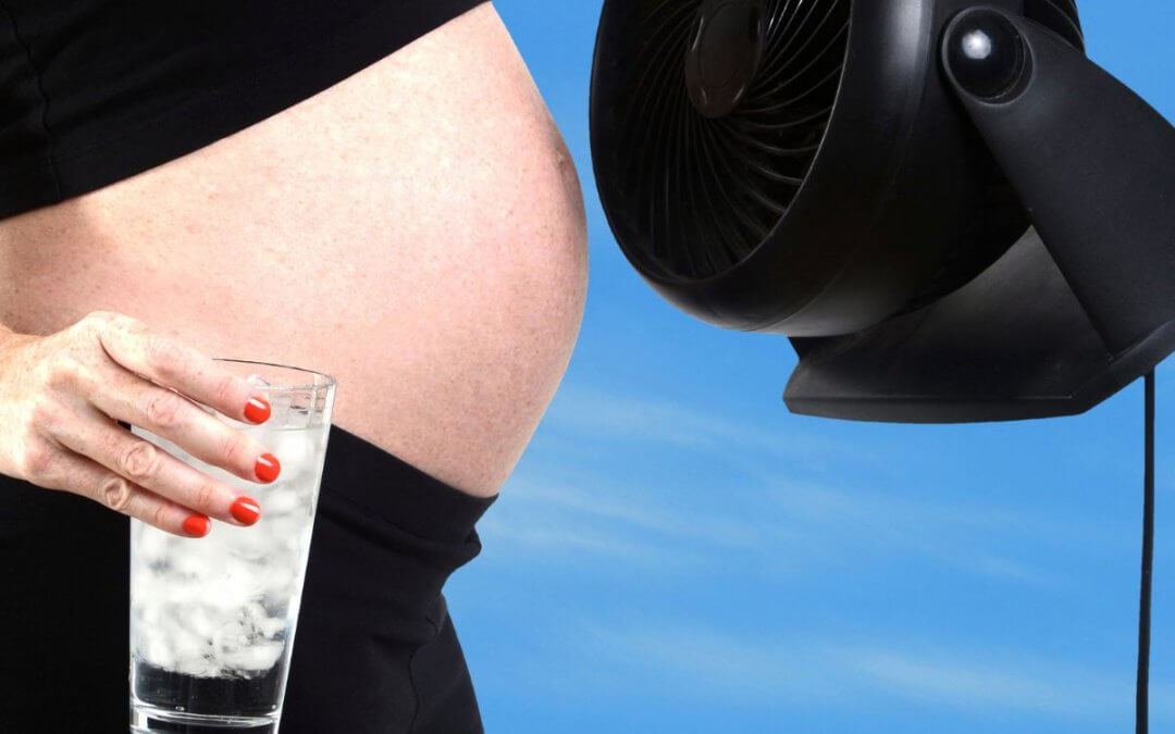 Pregnant woman in a black cropped top holding a glass of Iced water in one hand a a fan in the other cooling her stomach.